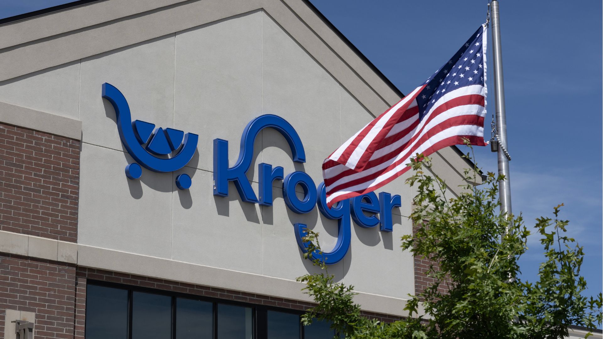 Blue Kroger supermarket sign on a store exterior with an American flag waving in the foreground.