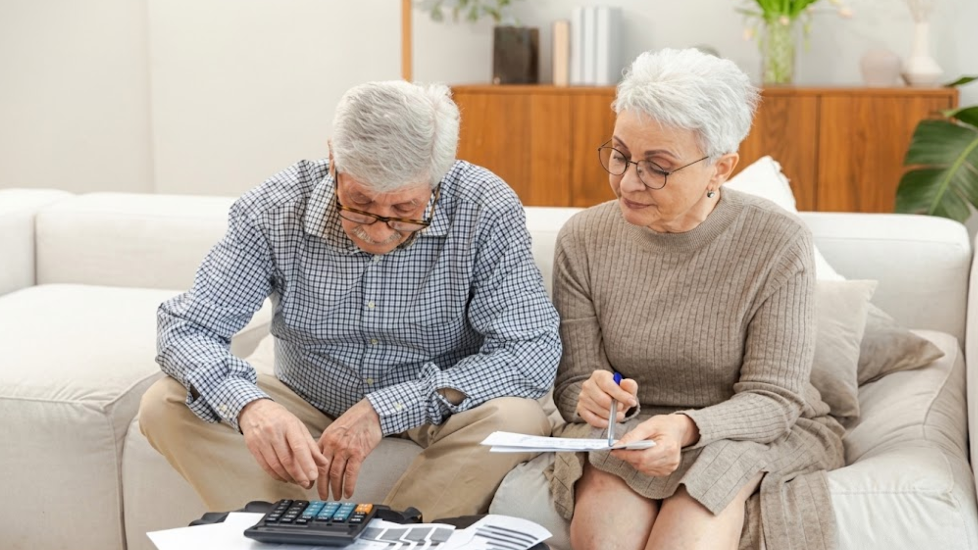 An elderly couple reviewing financial papers and using a calculator while sitting on a living room couch