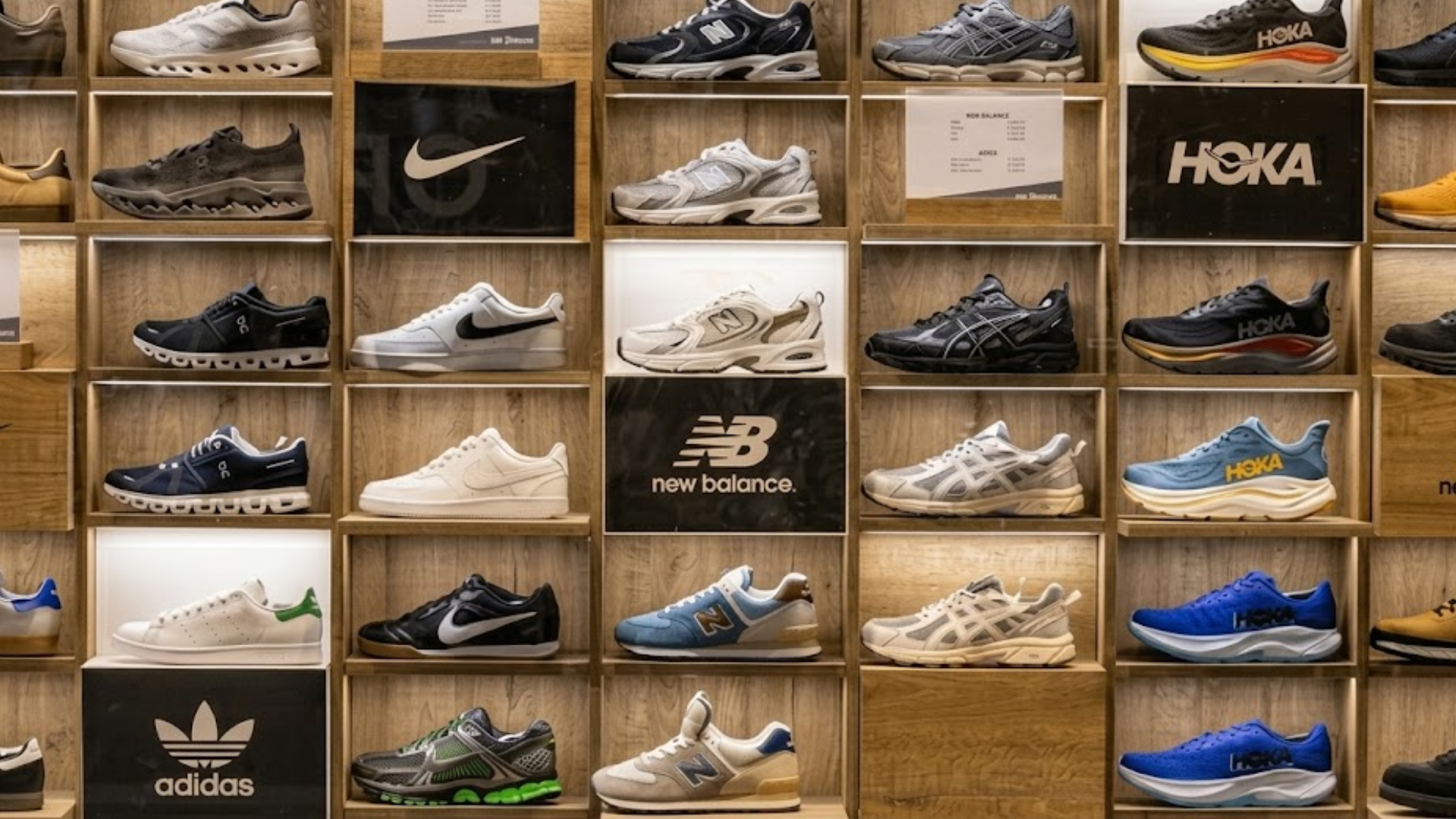 A brightly lit retail store display wall featuring neatly arranged rows of athletic sneakers and sports footwear from popular brands on wooden shelves.