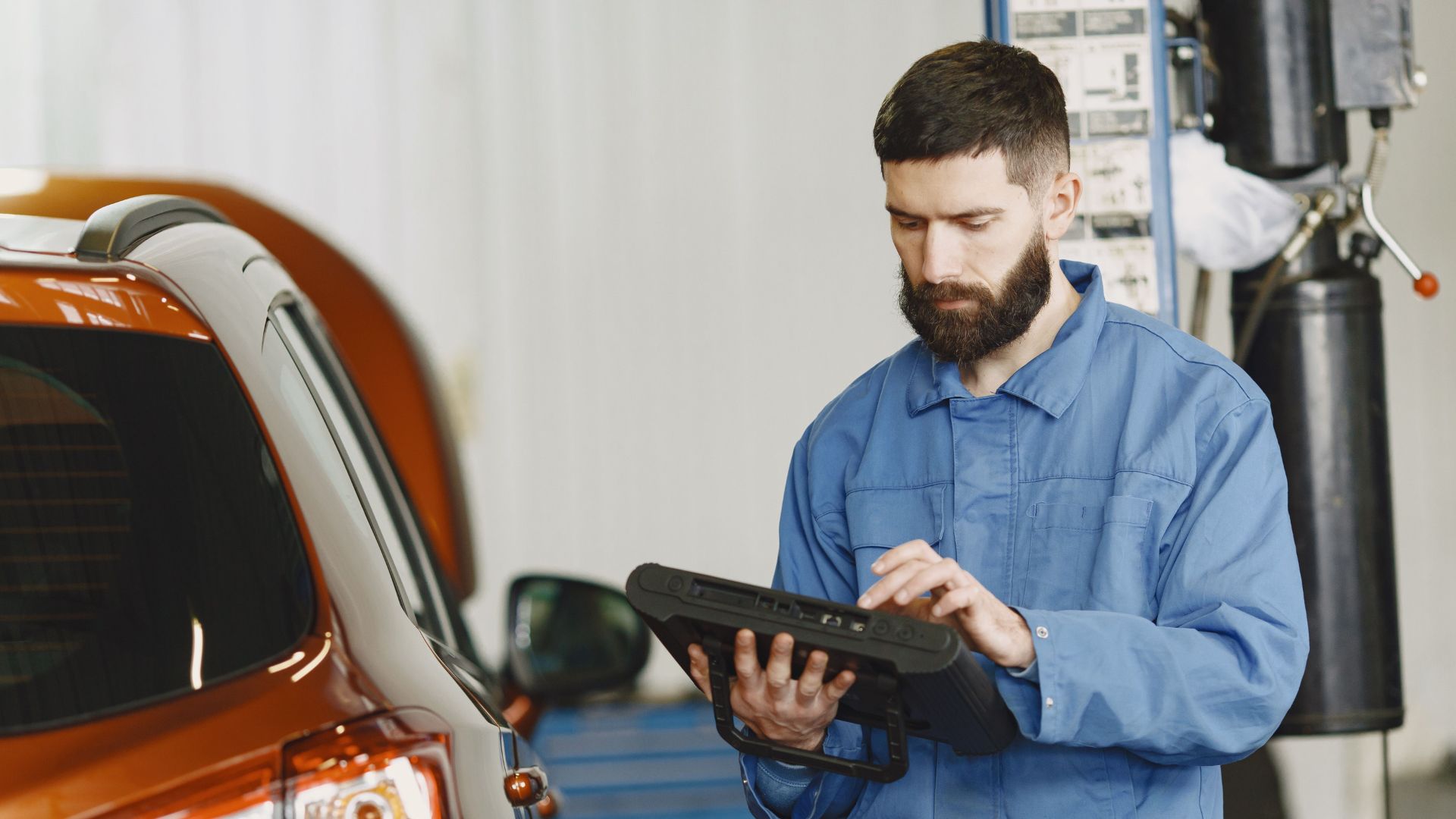A mechanic in blue coveralls using a tablet next to an orange SUV in a repair shop.