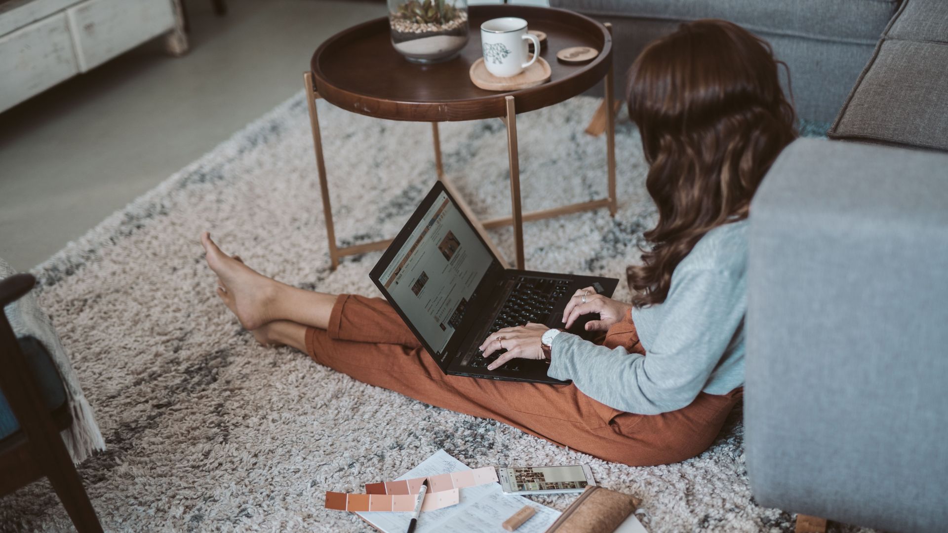 Woman sitting on a living room rug using a laptop beside a sofa and coffee table.