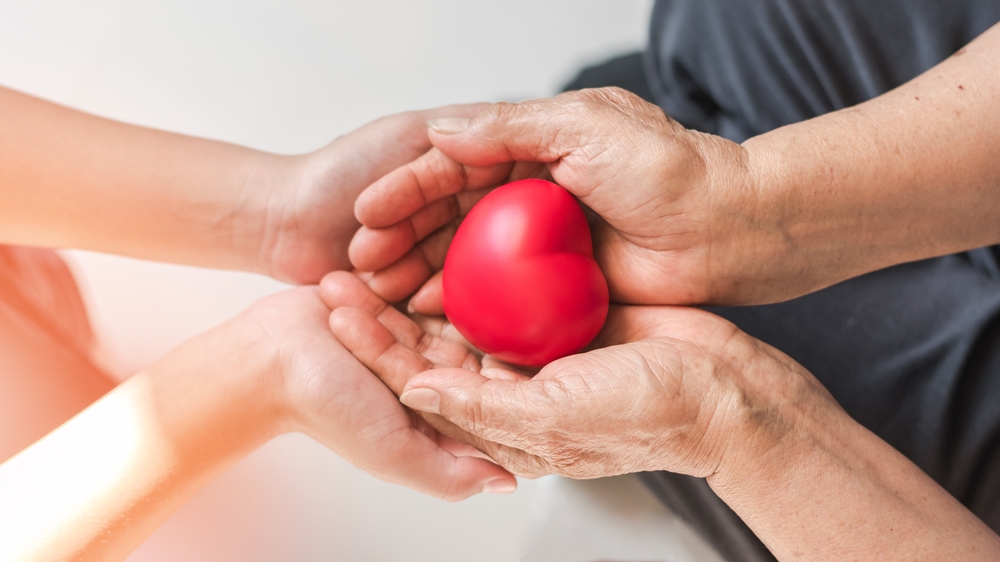 Older and younger hands holding a red heart together.